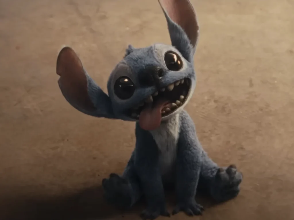 Imagem do personagem Stitch no live-action de 'Lilo & Stitch'. Ele aparece sentado no chão, com expressão animada, olhos grandes e brilhantes, orelhas erguidas e a língua de fora.